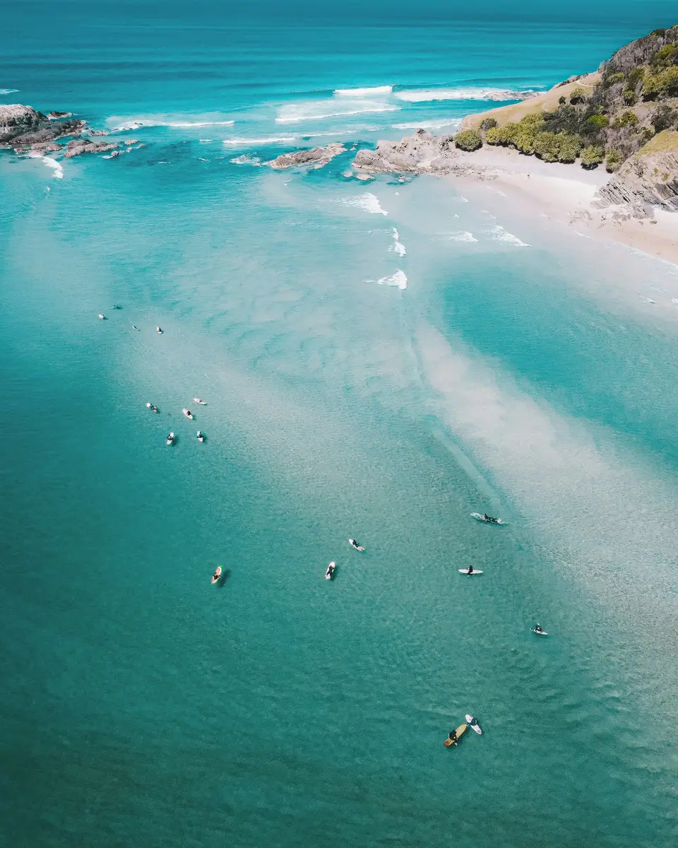 Aerial view of turquoise surf, sandy beach, rocky headland, and people in the water in Australia.