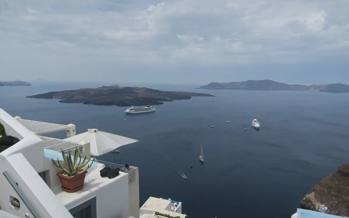 Whitewashed terrace above a broad Greek island bay with boats and low hills in the distance.