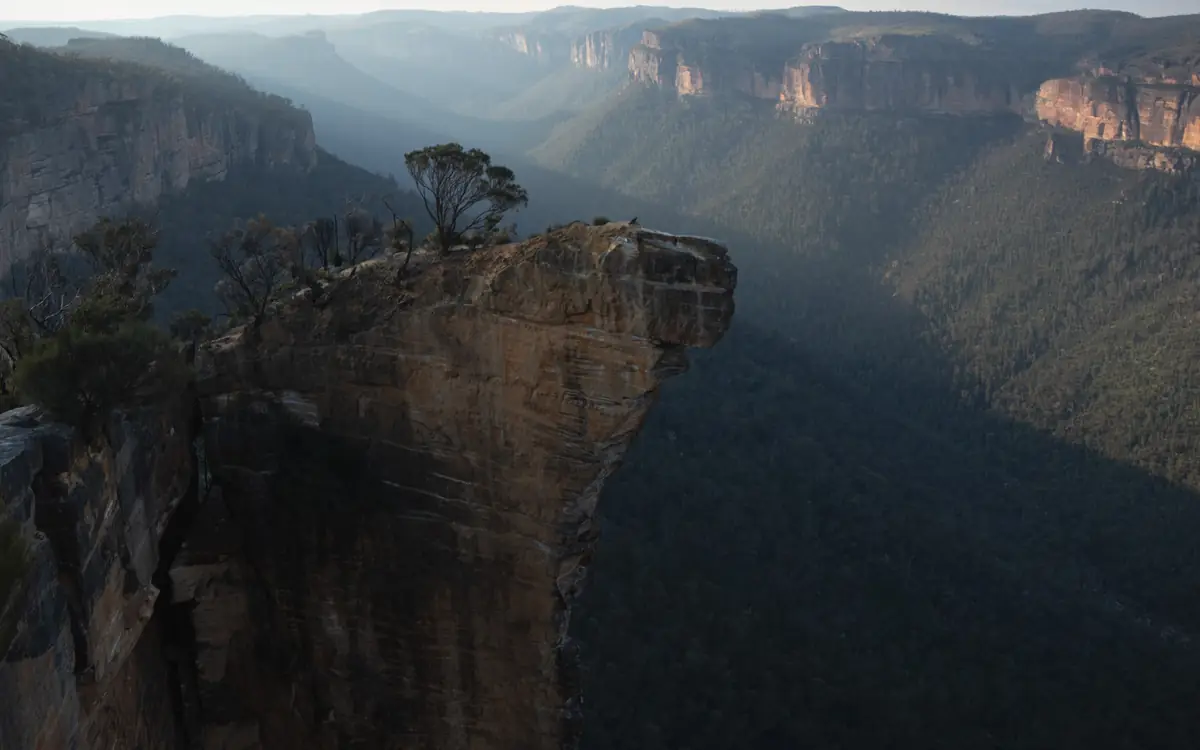 Sandstone cliff ledge above a deep forested valley and hazy escarpments in New South Wales.