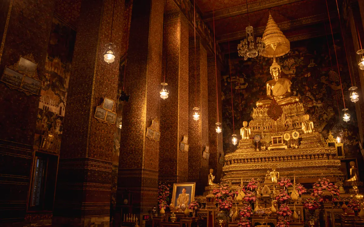 Golden seated Buddha statue inside a Bangkok temple hall with monks seated nearby.