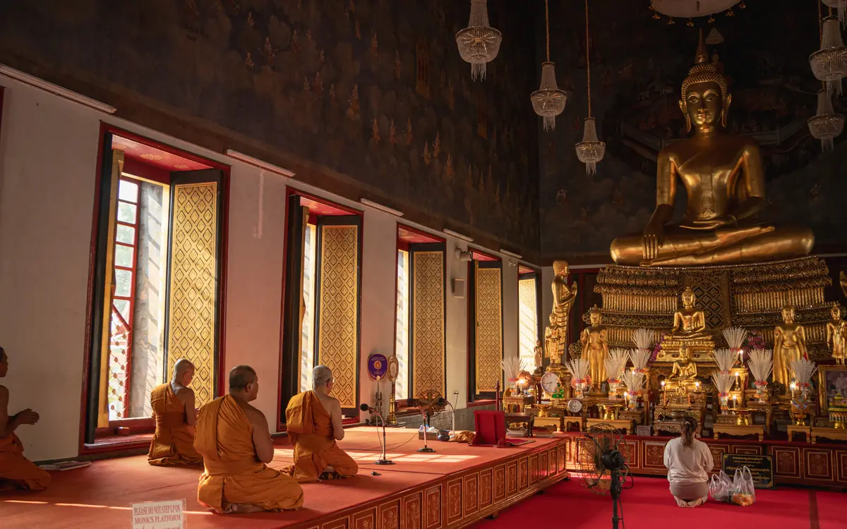Golden seated Buddha statue with monks and visitors inside a decorated temple hall in Bangkok.