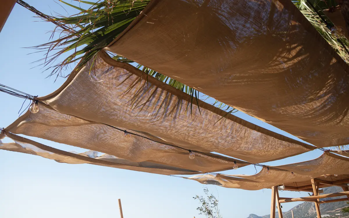 Woven shade cloths, palm fronds, and string lights against a bright sky on Crete.