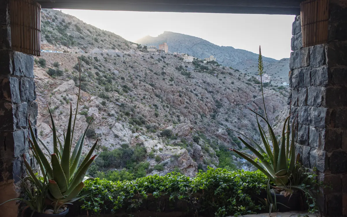 Stone-framed view of rocky slopes, sparse buildings, and agave-like plants in an Oman mountain village.