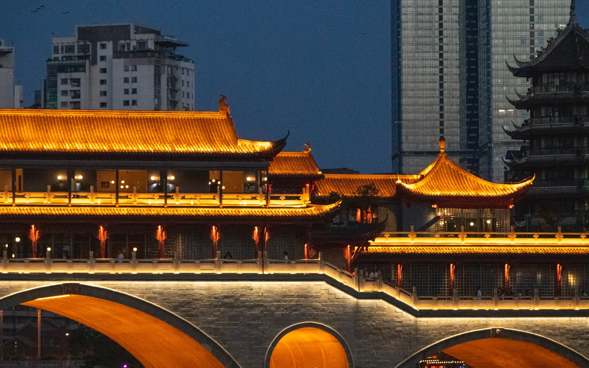 Illuminated traditional-style bridge reflected in water with high-rises behind it in Sichuan.