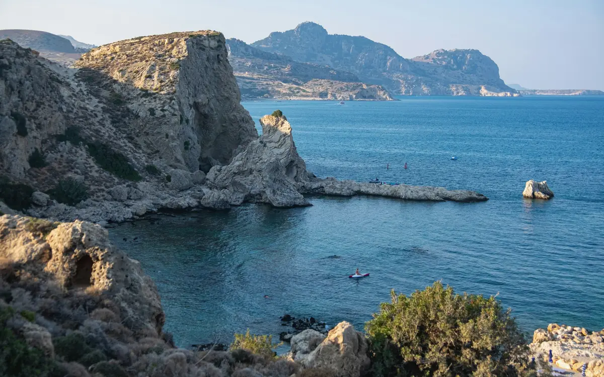 Rocky island bay in Rhodes with blue water, small boats, cliffs, and dry mountains.
