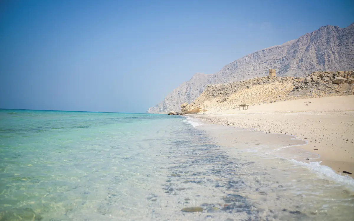 Turquoise water, pale sand, and a dry rocky mountain behind a beach in Oman.