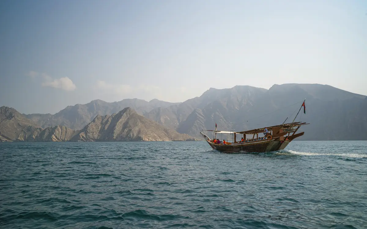 Traditional wooden dhow on blue water with steep dry coastal mountains in Oman.
