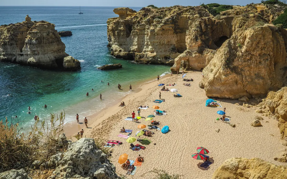 Small sandy beach cove with limestone cliffs, turquoise water, umbrellas, and people in the Algarve.