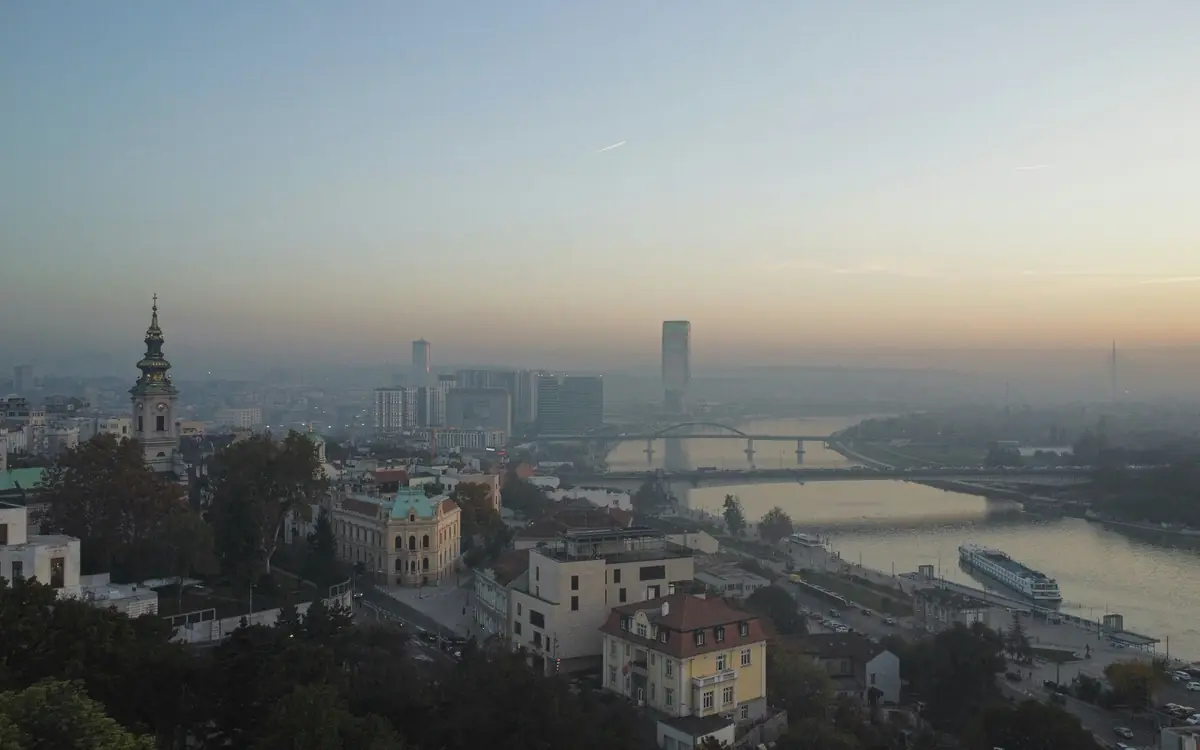 Belgrade city skyline along a river with a church tower, bridges, and modern towers in haze.