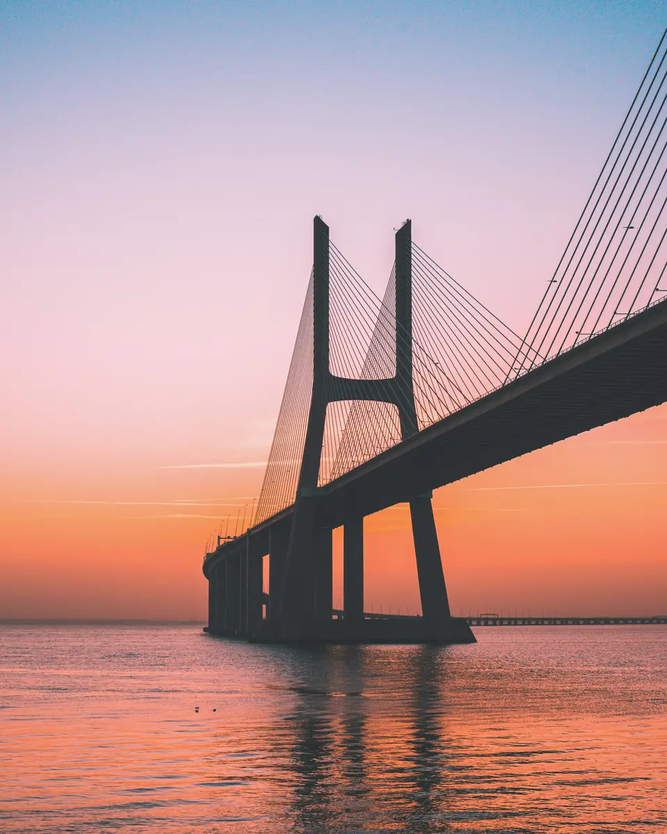 Red suspension bridge in silhouette over water at sunset in Portugal.
