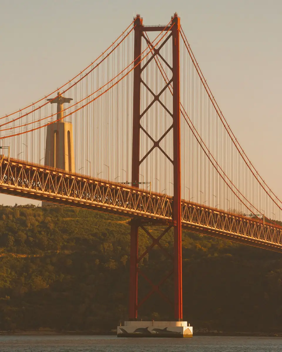 Red suspension bridge pylon above water in golden light in Portugal.