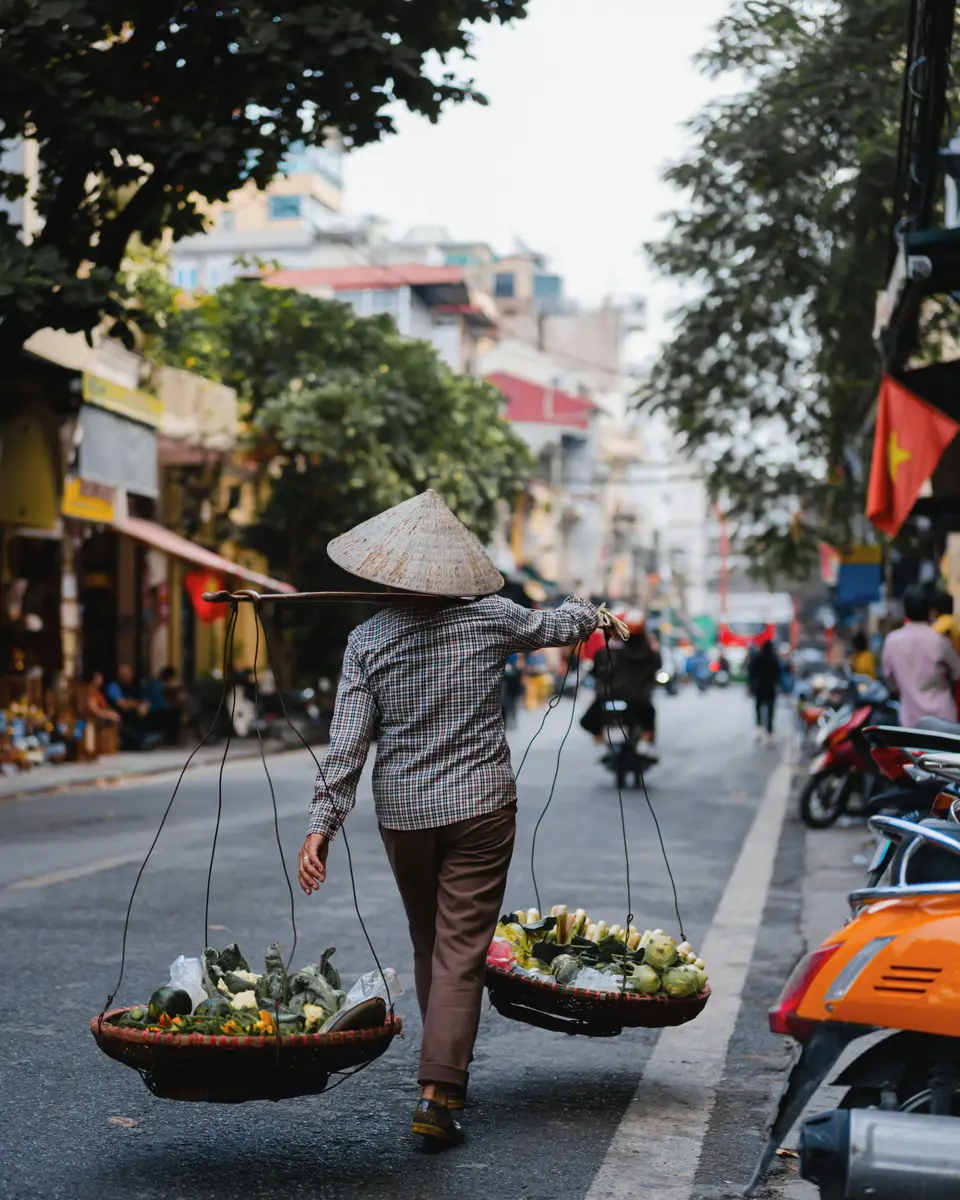Street vendor carrying baskets on an urban street with scooters and a red flag in Vietnam.